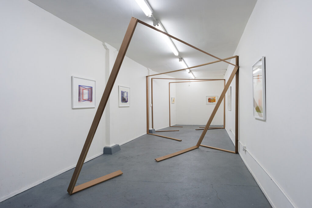 An art installation with a large wooden structure in the middle of a gallery. and works mounted on the walls.