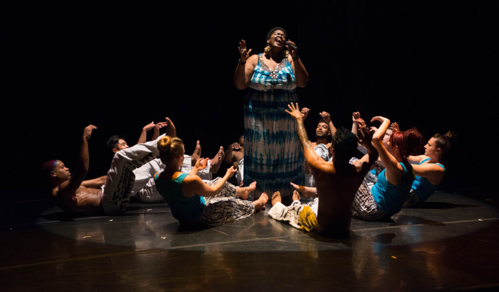 A person singing in the middle of a circle of people dancing on the floor around them.