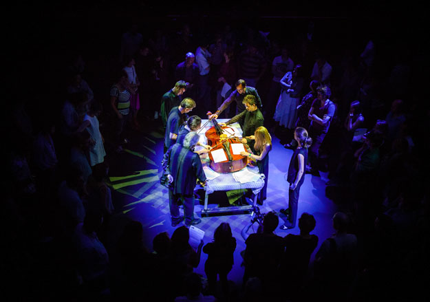 People grouped around a cello lying on a table.