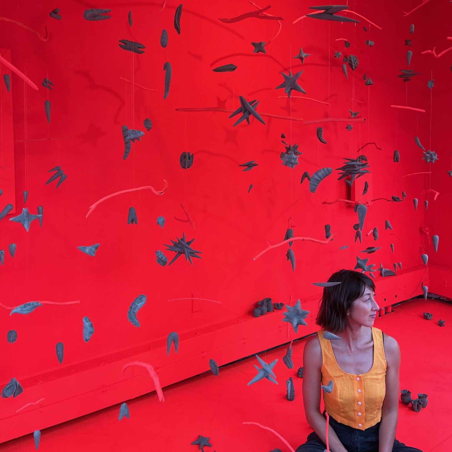 The artist in a red room with gray objects hanging from the celing around them.