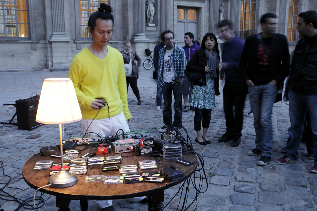 A person standing outside with cassette tapes and players on a table with a group of people looking on.