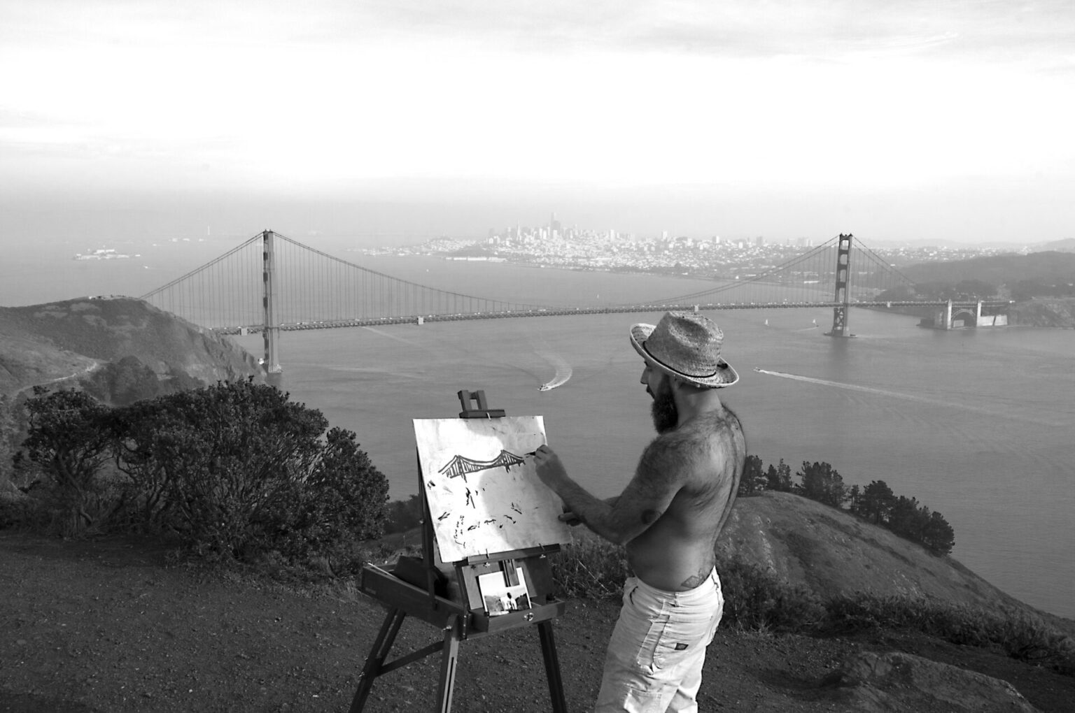 José in shorts and a fedora making a painting while standing on the cliffs of the Marin Headlands in front of the Golden Gate Bridge.
