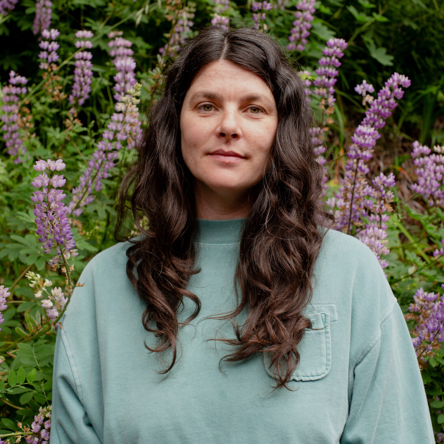 Portrait of Nicole wearing a blueish green long sleeve while standing in front of purple flowers.
