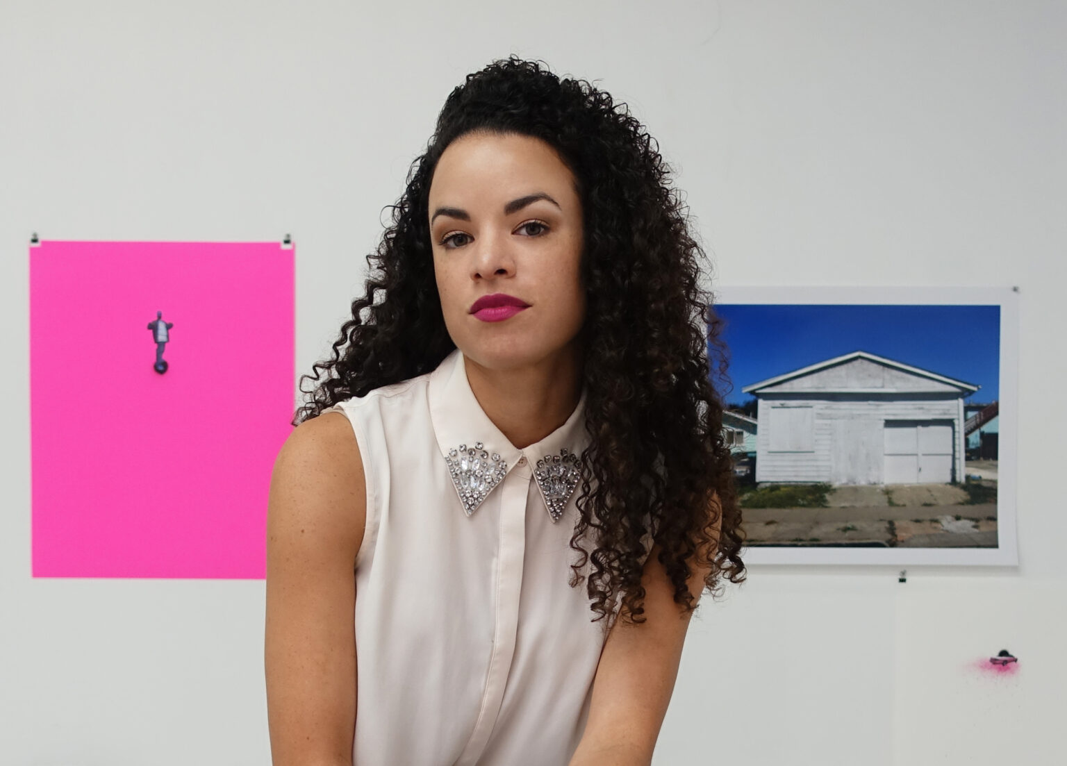 A portrait of a person in front of artworks mounted on a white wall.