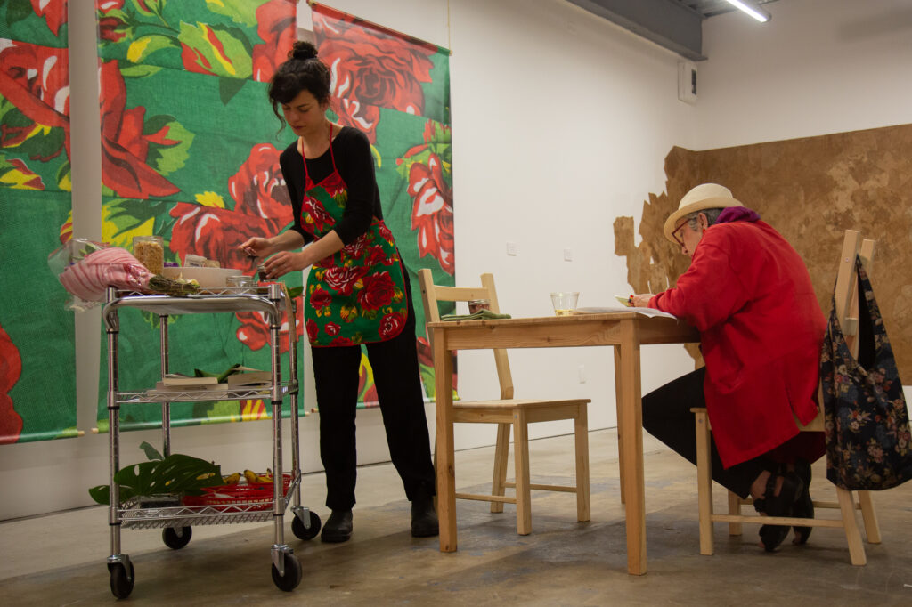 A person working at a rolling cart next to a person sitting at a table in an art gallery.