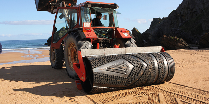 A piece of heavy machinery using a large roller to imprint a pattern into sand.