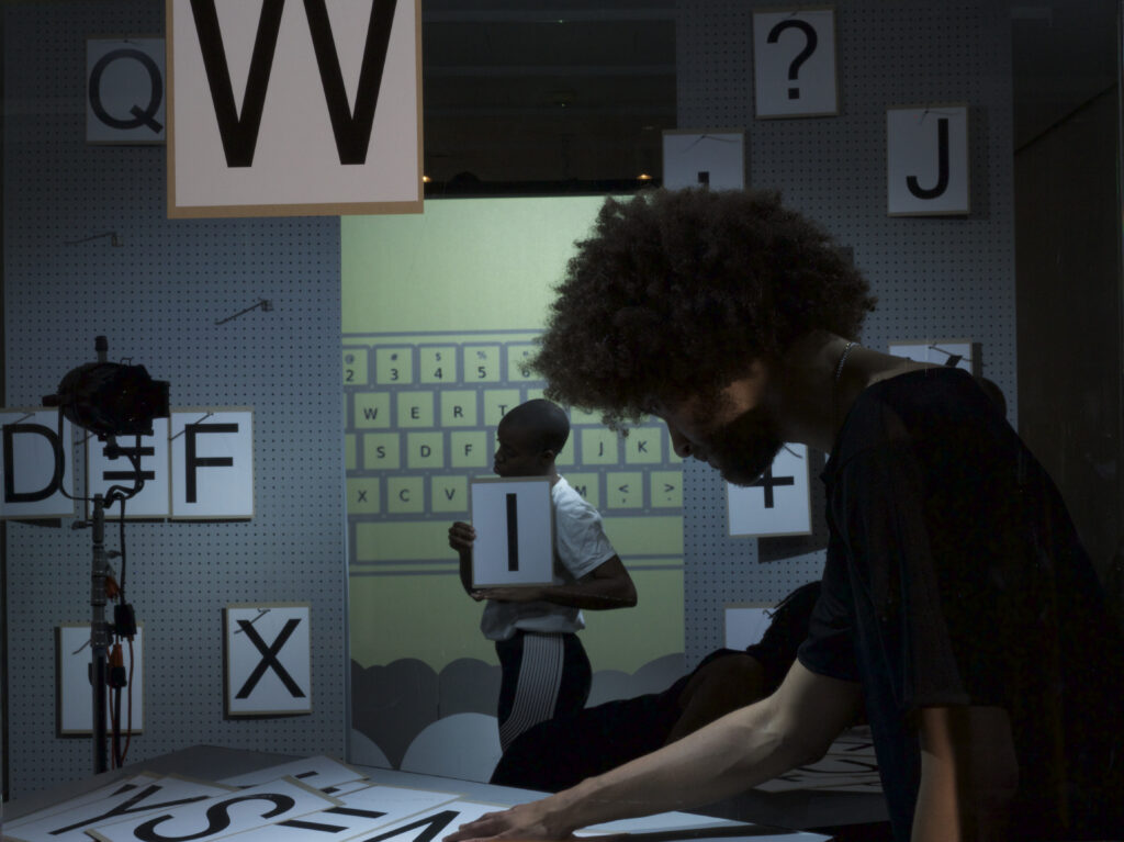 A person looks through printed paper letters in the foreground while another person holds up a letter in the background.