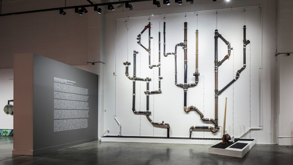 An image of an art installation comprised of pipes and plumbing fixtures mounted to a gallery wall.