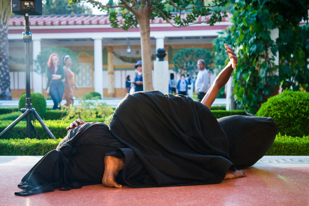 A person draped in dark fabric while holding an expressive pose.