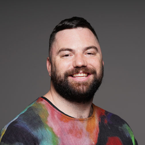 A portrait of a person in a multi-coloured shirt in front of a grey background.