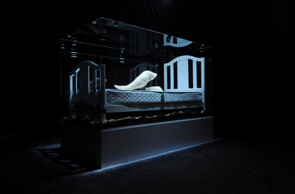 Shadowy image of an art installation including a bed frame and sculptures within a glass vitrine.