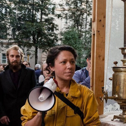 A person holding a megaphone next a samovar and tea glasses.