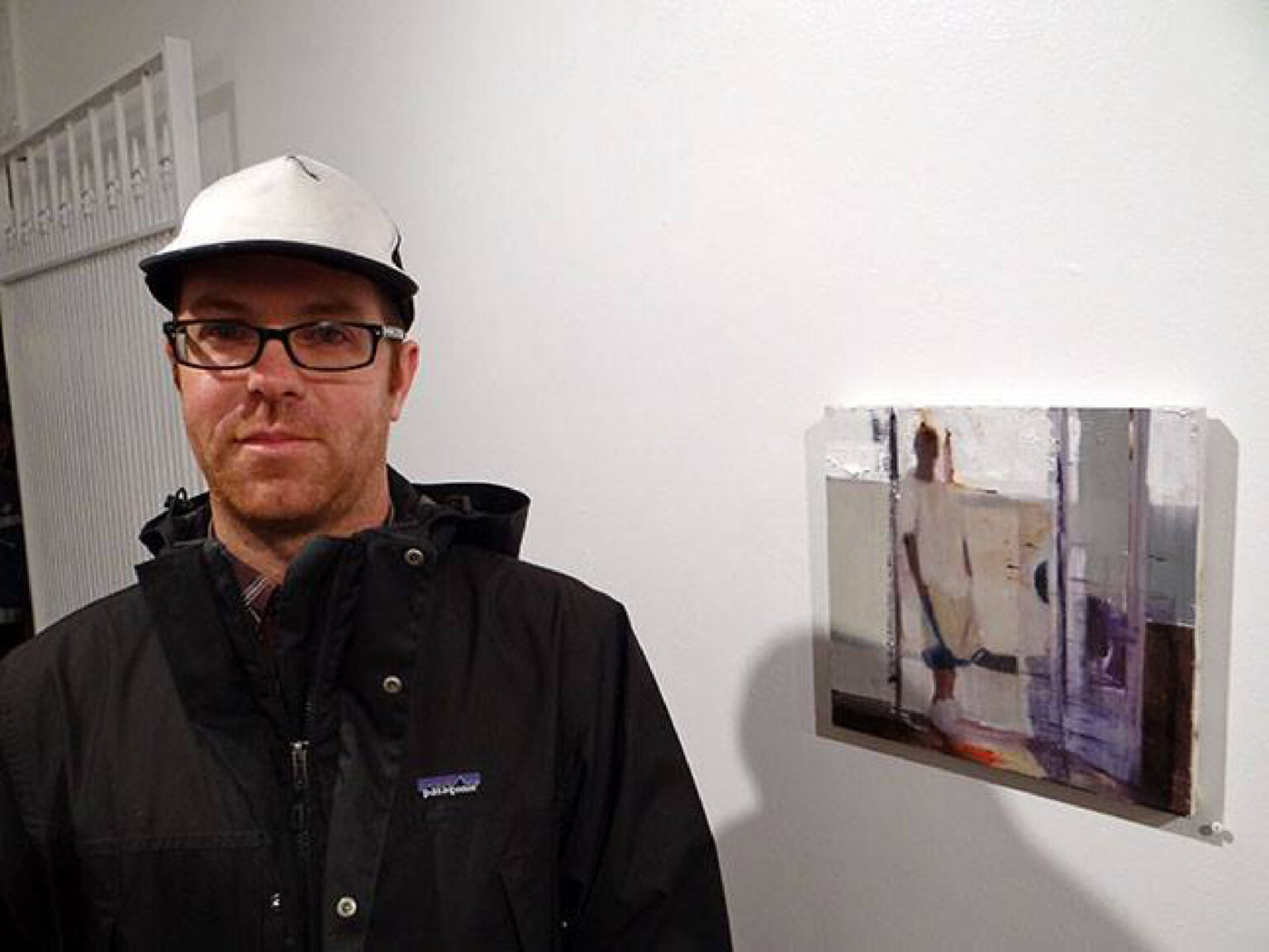A person in a hat standing next to painting on wall.