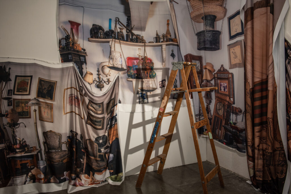 An installation comprised of a ladder, printed fabric, framed works, and other items in a white-walled room.