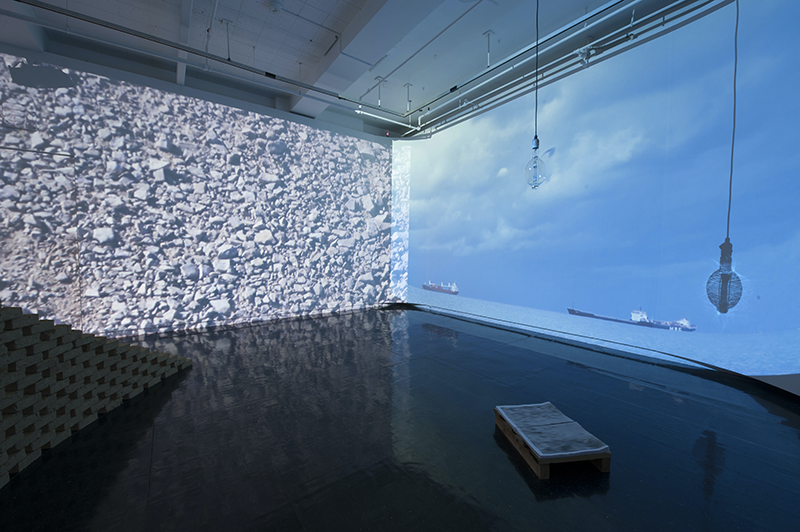 Room with video projections on the walls: on the right an ocean and clouds, on the left, rocks. A lightbulb, stacked bricks, and stack of papers sit in the room.