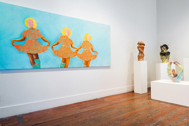 Gallery with white walls and wood floors, with a painting on the left wall depicting three figures curtsying against a blue background, and sculptures on pedestals at the far end of the room