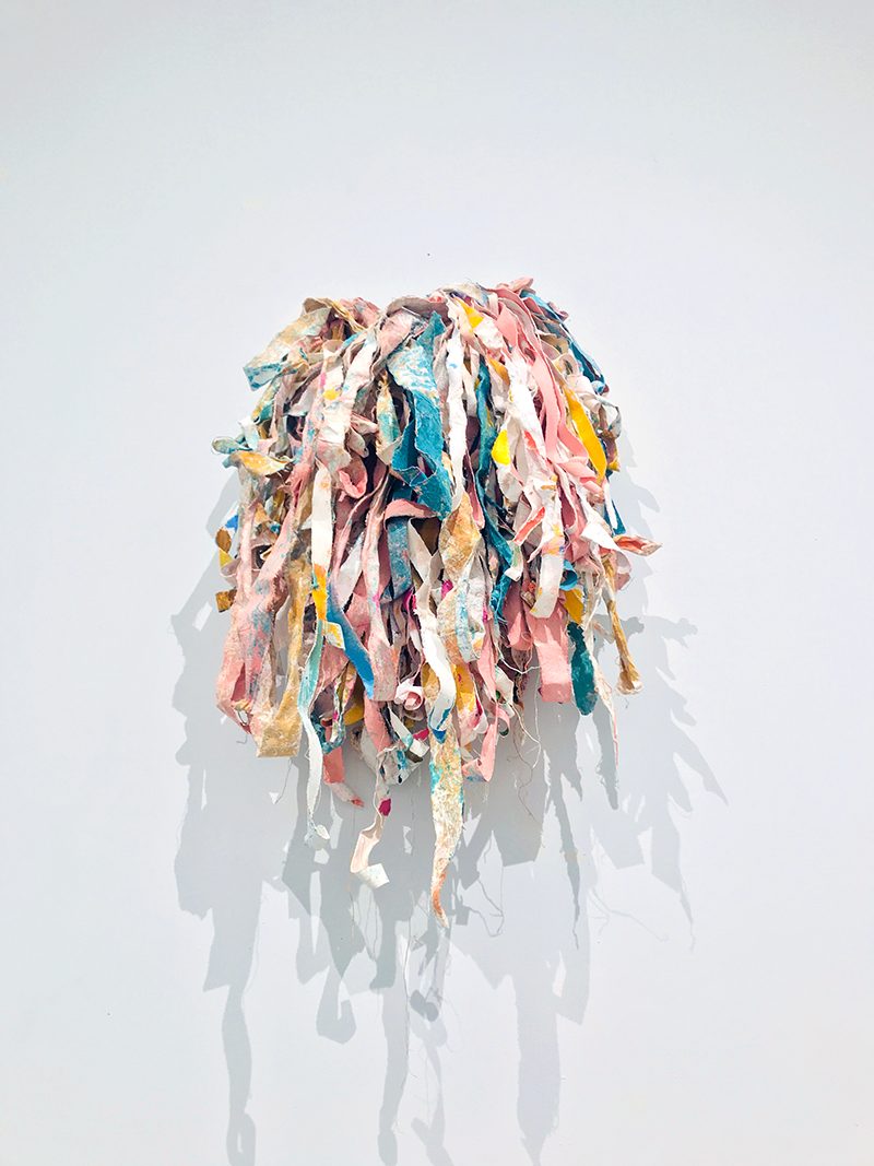 Colorful sculptural form comprised of strips of cloth formed around a ball or central form, mounted on the wall.
