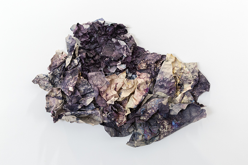 an object of dark purple and blue dyed paper, with some bleached pale portions, crumpled and set atop a reflective, mirrored surface
