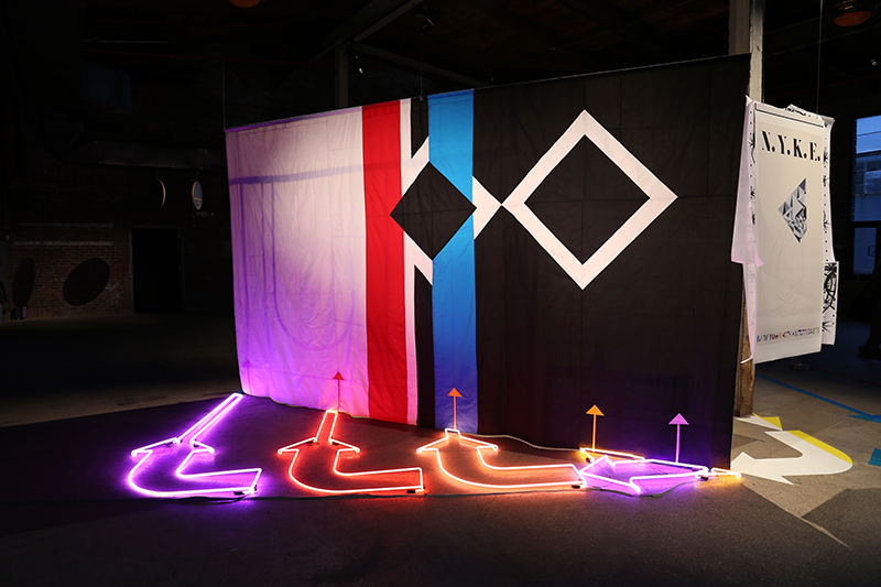 An installation in a dark gallery featuring a quilt with large black and white color blocks and red and blue vertical color bands hanging suspended in the space, with neon lights in the shape of arrows on the floor