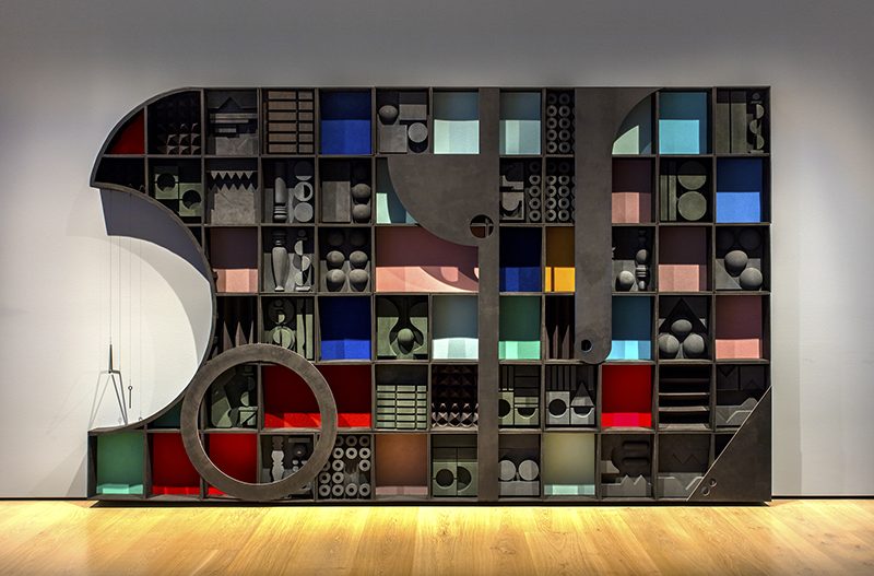 A sculpture comprised of shelving-unit-like blocks painted black or dark grey, some interiors painted with various jewel tones, and with abstract shapes inside. One end of the structure has a curved section excised, and a metal musical triangle hangs there.