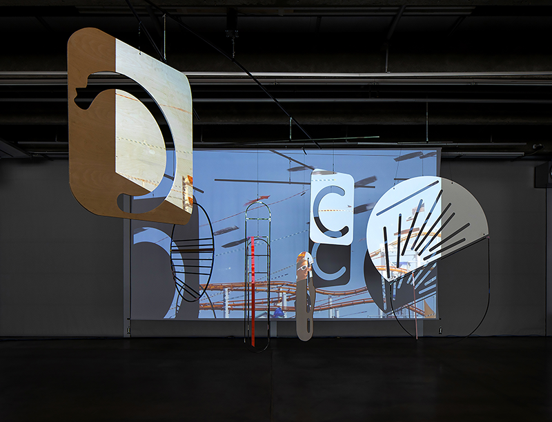 A projection on a wall of blue skies is obscured in part by hanging abstract shapes made out of wood, which cast shadows on the projection