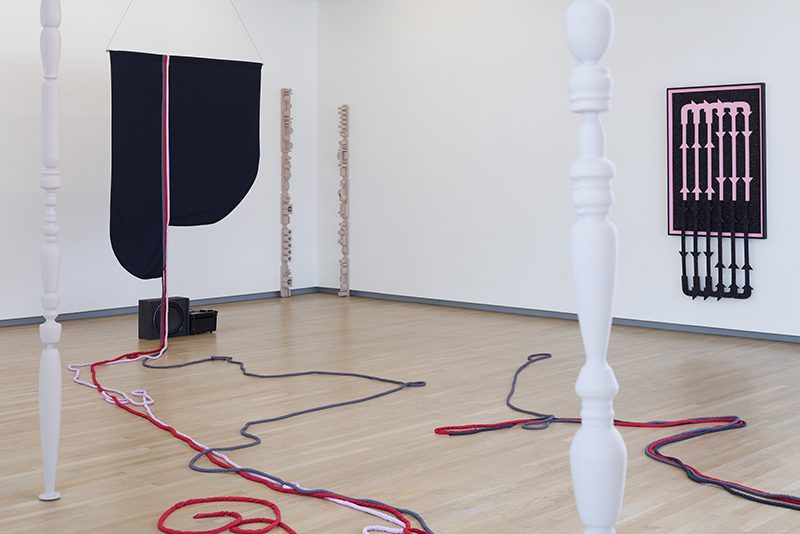 a gallery with white walls and wood floors, containing a set of objects including painted wood lathed forms in the foreground, ropes or cords in red and black strung across the ground, and large black and pink forms hanging on far walls