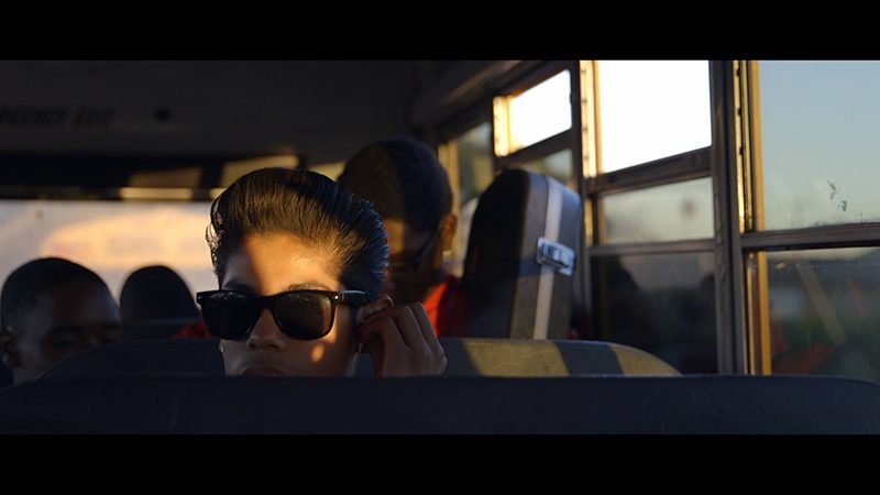 A person on a bus sits low in a seat, only part of their face, obscured by large black sunglasses, is visible. Behind them are musical instrument cases.