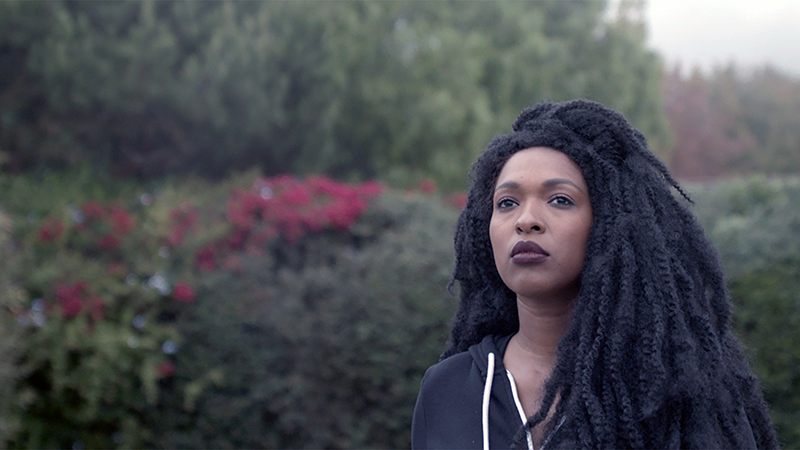 A woman with dark skin and black har, wearing a black hoodie and dark lipstick, stands outside. Behind her are hedges with red flowers.