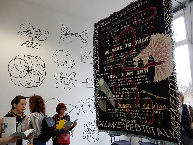 A large woven textile with text and images hangs in a gallery type space. Diagrams are visible on the wall in the background, and people stand in the space.