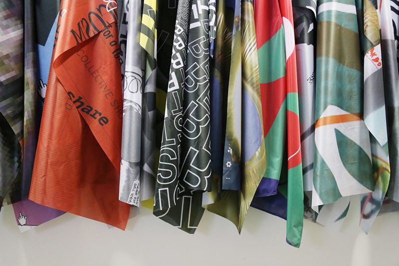 A series of silk scarves in multiple colors hang in front of a white wall