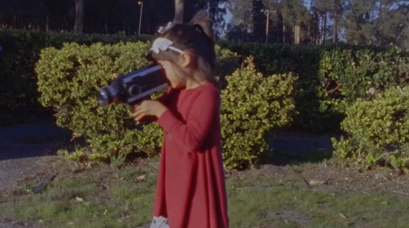 A young peson in a red dress and hair band holds a handheld film camera to their eye.