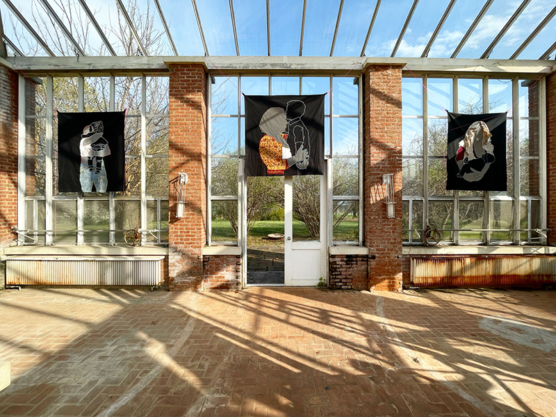 Textile images of abstracted figures on a black ground hang on display in an open, airy, brick and glass space.