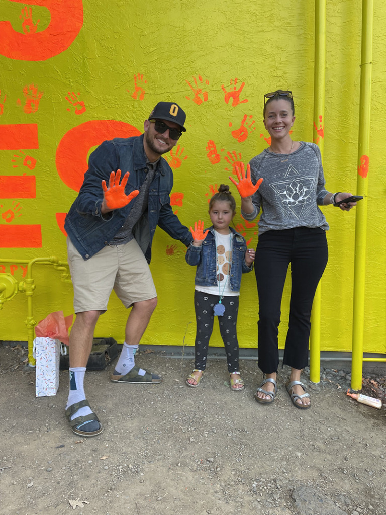 A family standing in front of a bright yellow wall with orange handprints, their hands covered in bright orange paint.