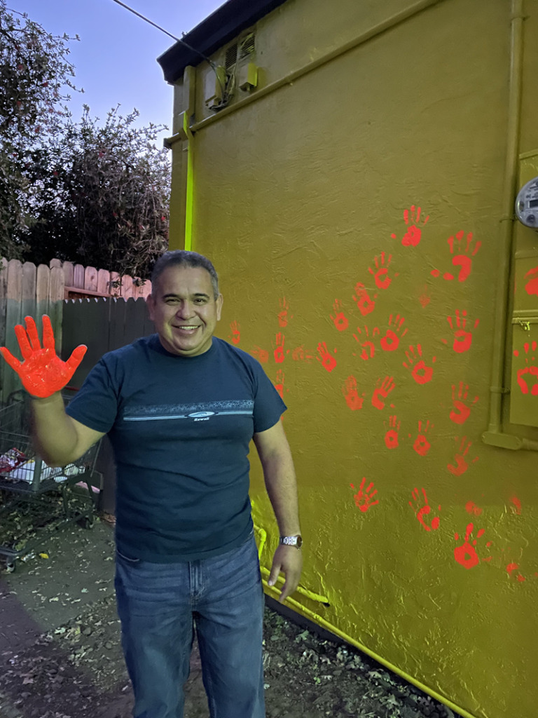 A person standing in front of a bright yellow wall with orange handprints, their hands covered in bright orange paint.