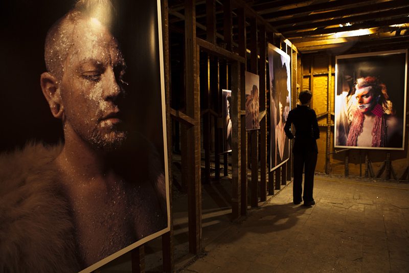 Photographic portraits hung in a dimly lit building that appears to be under construction. A person stands viewing a photograph.
