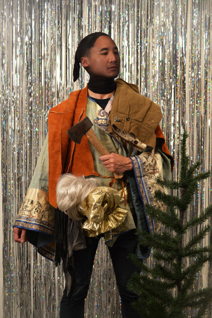 Portrait of a person in an elaborate and haphazard costume, holding a small hatchet and standing beside a small artificial pine tree in front of a glittery backdrop.