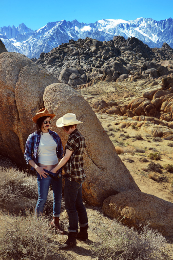 Two people in cosyume including blue jeans, flannel shirts, sunglasses, and cowboy hats in a landscape of large boulders and desert scrub. One person stands facing the other with their hand down the front of the pants of the other.