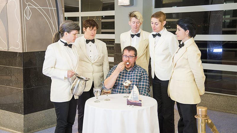 A white man in a plaid shirt sits at a table with a white table cloth, eating an ice cream from a tall glass. He is surrounded by people dressed in costume as servers in white tuxedo shirts and ties.