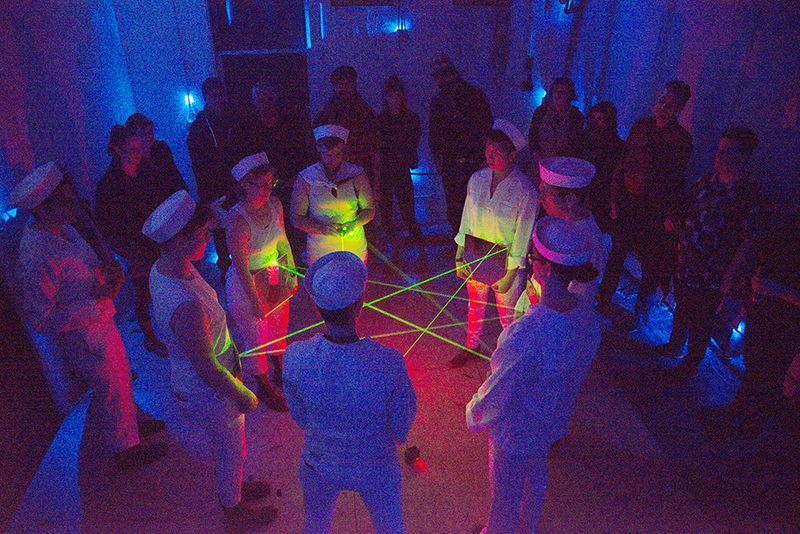 A group of people in sailor garb standing in a circle, with beams of yellow-green light between them.