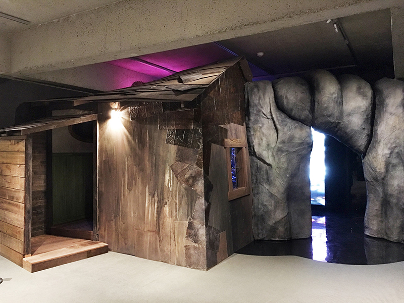 An installation resembling a weathered, wooden shack and boulders.