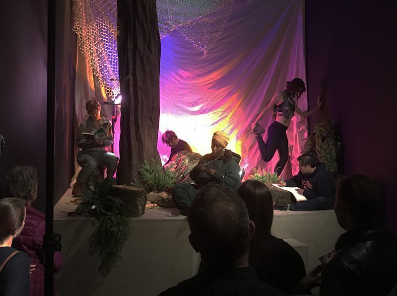 Various people sitting, standing, and lying in a space lit with purple and pink lighting.
