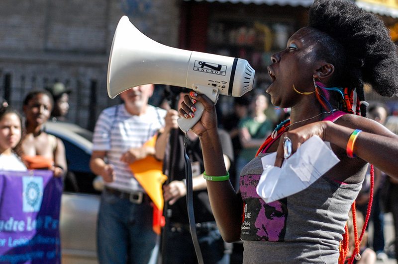 Photograph of a Black person shouting into a megaphone before a crowd of people.