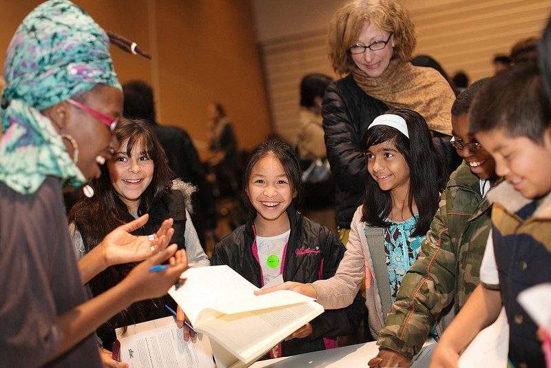 A Black person speaking with a multi-ethnic group of children, who are holding books out.