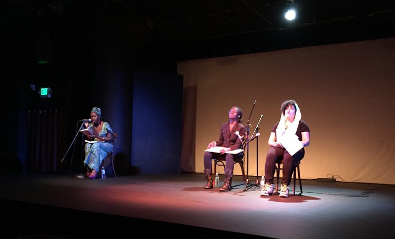 Three Black people on a stage, sitting in chairs before microphones.