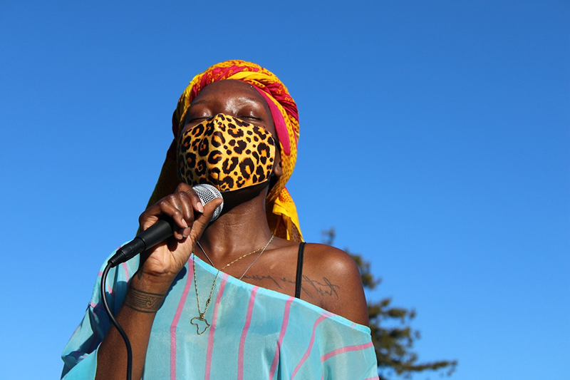 Photo of a Black person in a blue garment, red and yellow headscarf, and yellow and black face-mask holding a microphone against a blue sky.