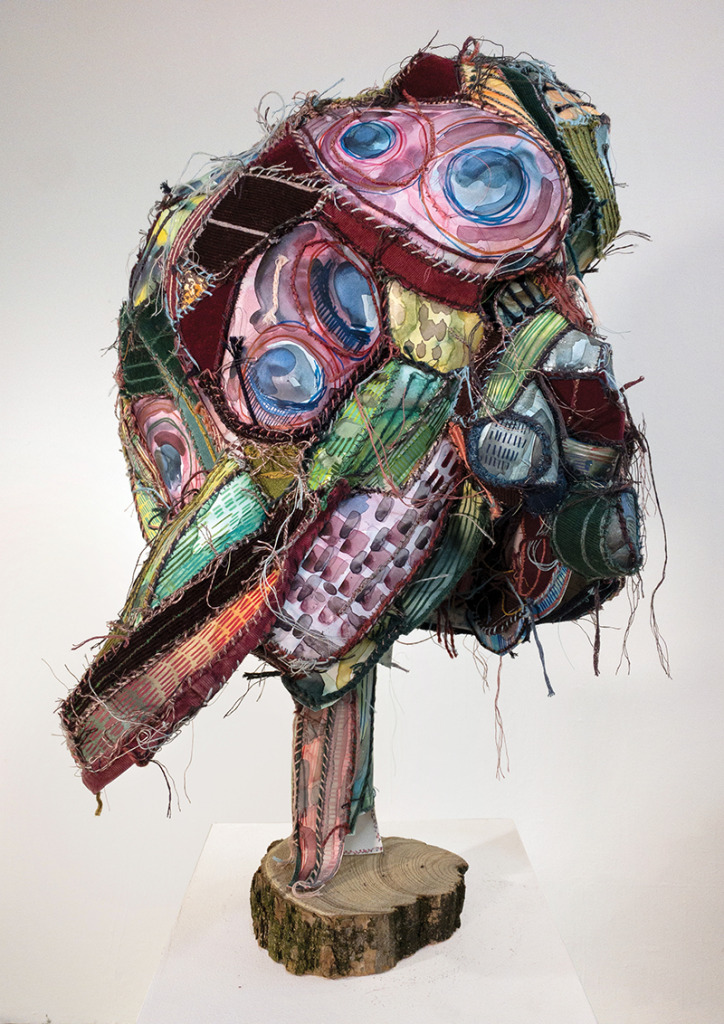 A colorful sculpture of stitched together textile mounted on a wood base.