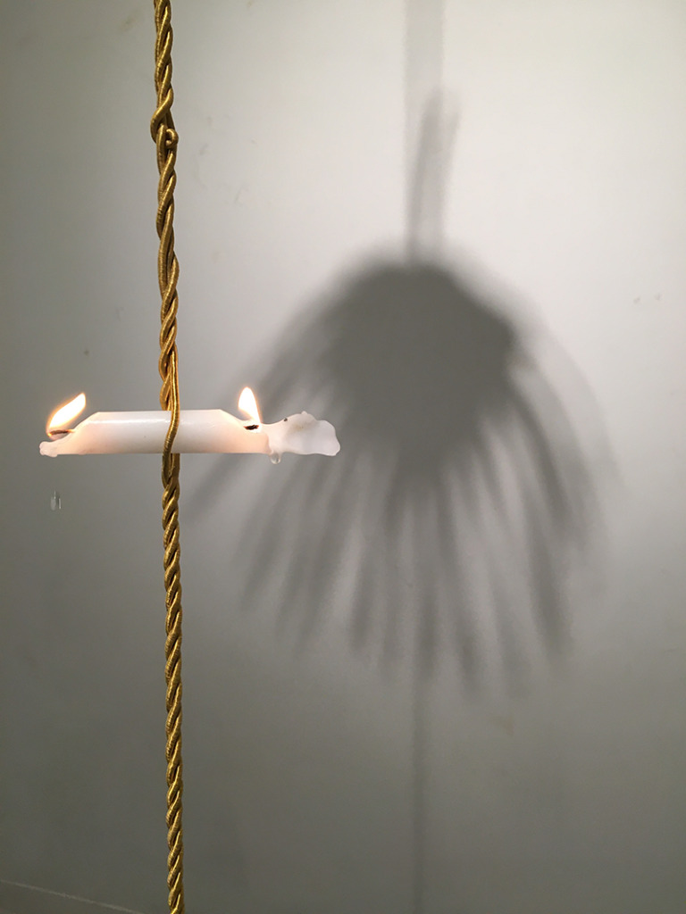 A candle burning on both ends, held horizontally aloft by a gold cord.