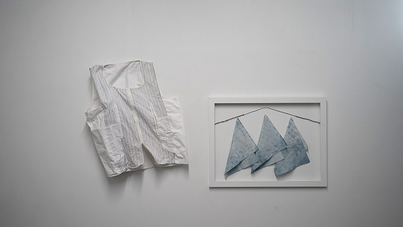 A white vest hung on a white wall beside a white-framed set of three pale blue swatched of folded cloth.
