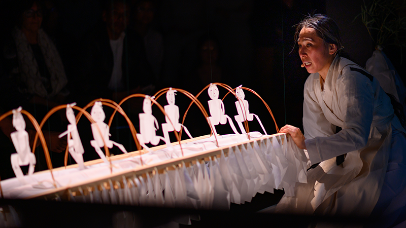 A sculpture of paper with arches supporting cut out figures is held and manipulated by a person in a white robe