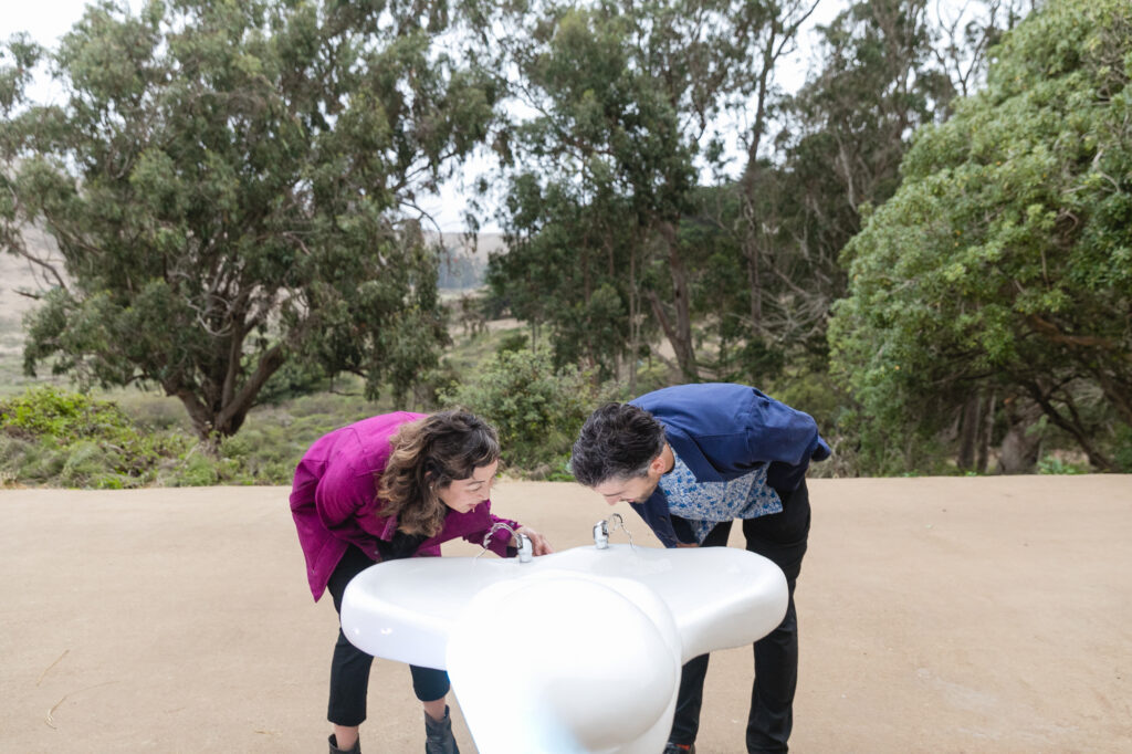 Two people drinking from the lumpy drinking fountain.